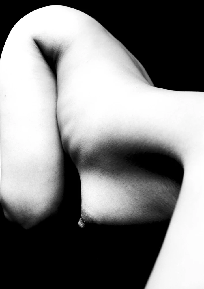 Abstract black and white study of a female torso emerging from darkness, its sculptural curves illuminated against a black background.