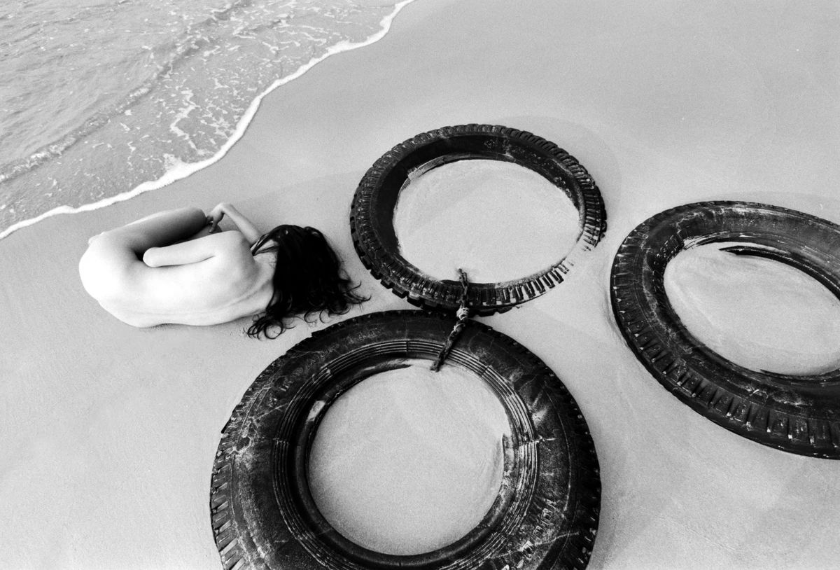 Black and white fine art nude photograph of a curled female figure on a beach among discarded tyres, with waves approaching the shoreline.