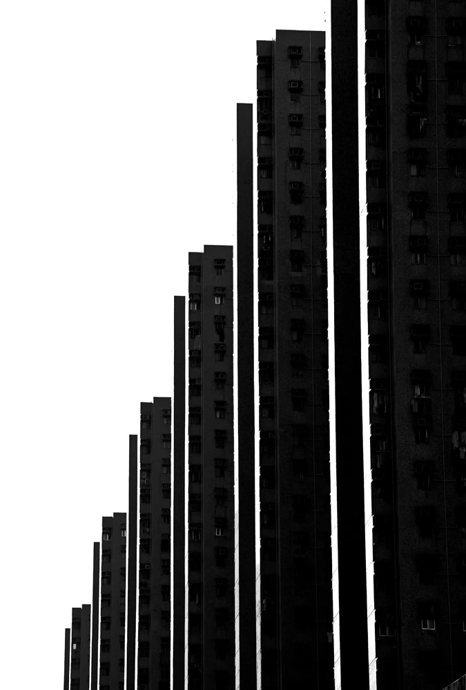 Black and white photograph of towering apartment buildings, creating a rhythmic pattern of vertical lines against an empty sky.